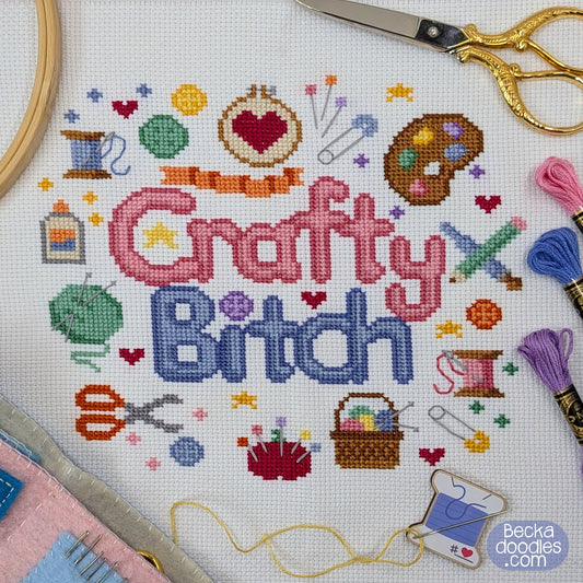 Cross-stitch design on white Aida fabric featuring the words "Crafty Bitch" in pink and blue thread, surrounded stitched crafting supplies and real sewing supplies.