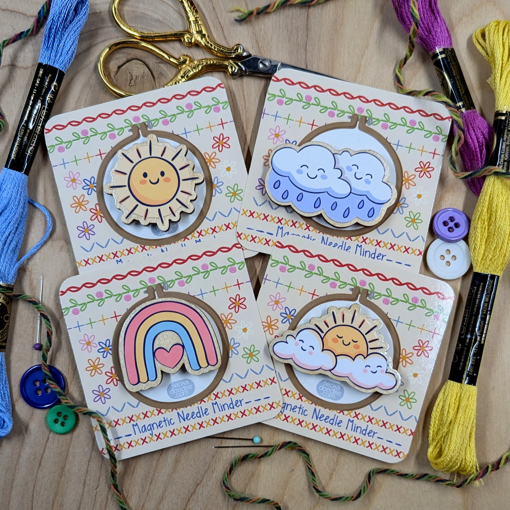 Set of colorful magnetic needle minders with sun, cloud, and rainbow designs on a wooden surface with embroidery tools.
