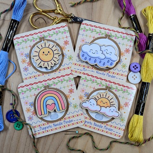 Set of colorful magnetic needle minders with sun, cloud, and rainbow designs on a wooden surface with embroidery tools.