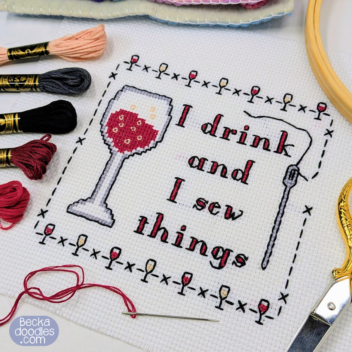 Completed cross-stitch design on white Aida fabric, featuring a wine glass and the words 'I drink and I sew things' surrounded by a stitched mini wine glasses border. Embroidery floss, a wooden hoop, scissors, threaded needle, and skeins of floss surround the design.