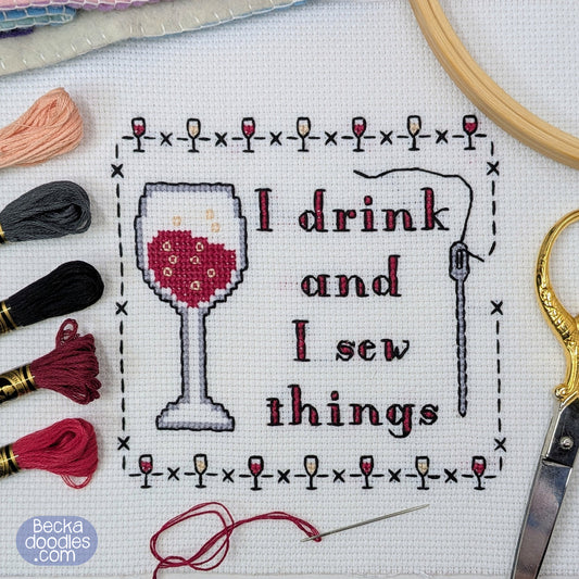 Completed cross-stitch design on white Aida fabric, featuring a wine glass and the words 'I drink and I sew things' surrounded by a stitched mini wine glasses border. Embroidery floss, a wooden hoop, scissors, threaded needle, and skeins of floss surround the design.