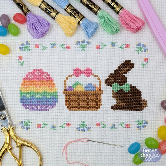 Cross-stitch design featuring a pastel striped Easter egg, brown basket filled with colorful eggs, and chocolate bunny with a green bow tie on white Aida fabric. Finished design is surrounded by sewing supplies and candy jelly beans..