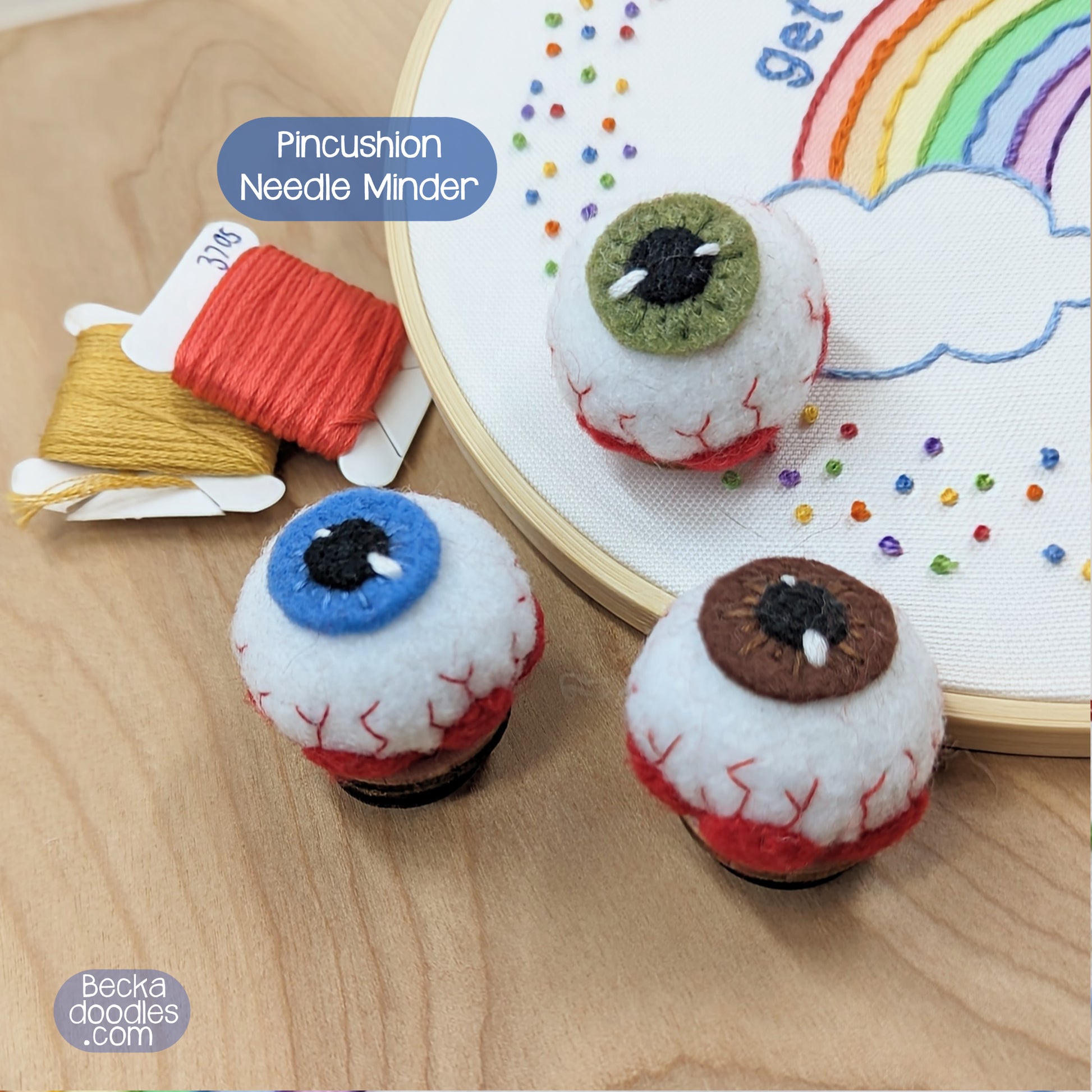 Eyeball-shaped pincushions with a rainbow and cloud embroidery design in the background.