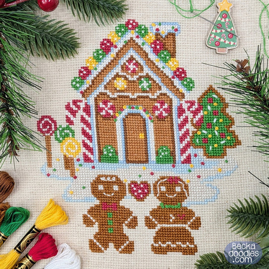 Holiday cross-stitch design of a gingerbread house with two gingerbread people, surrounded by pine branches, embroidery floss, and holiday decorations.