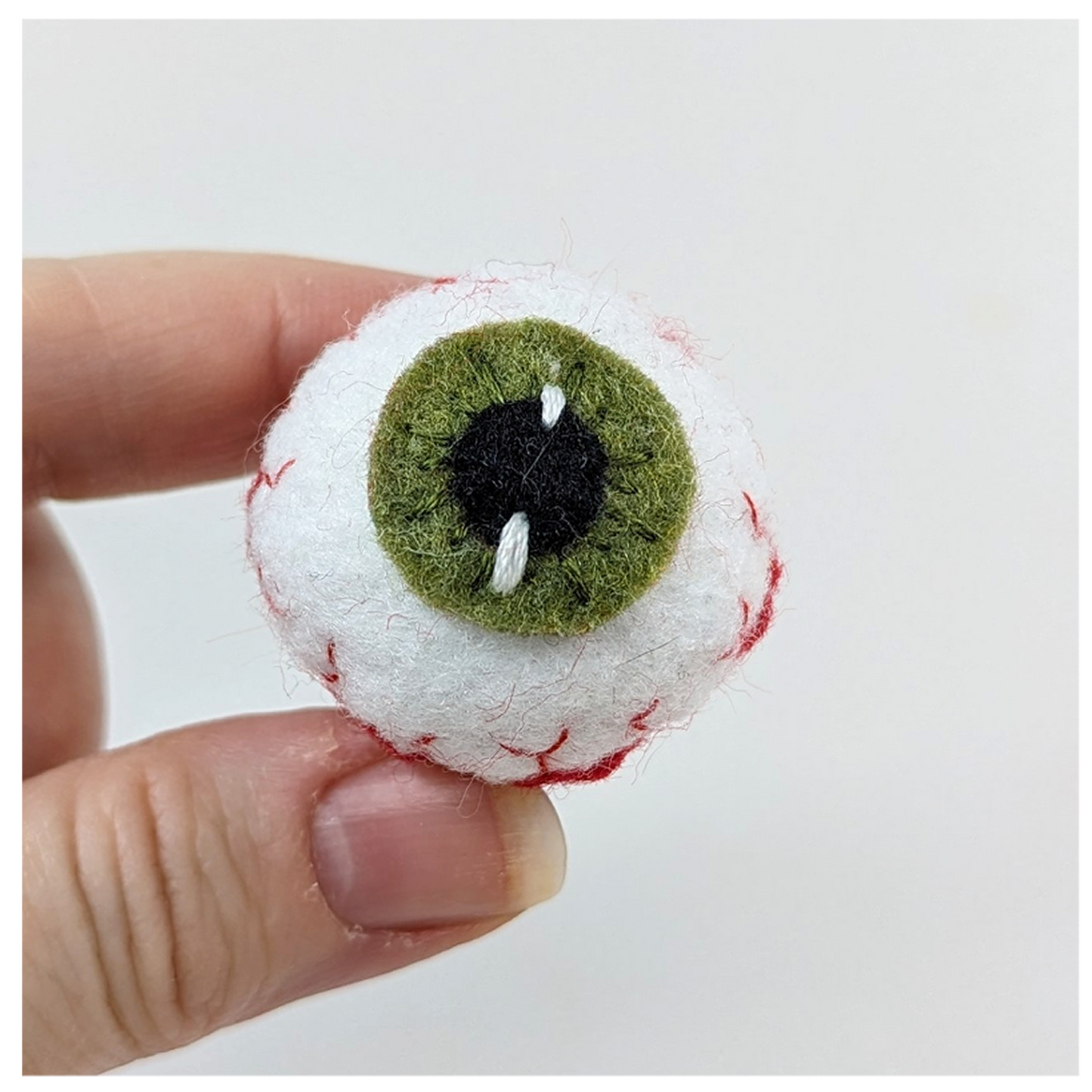 Hand holding a small felted eye with green iris and red sclera on a white background