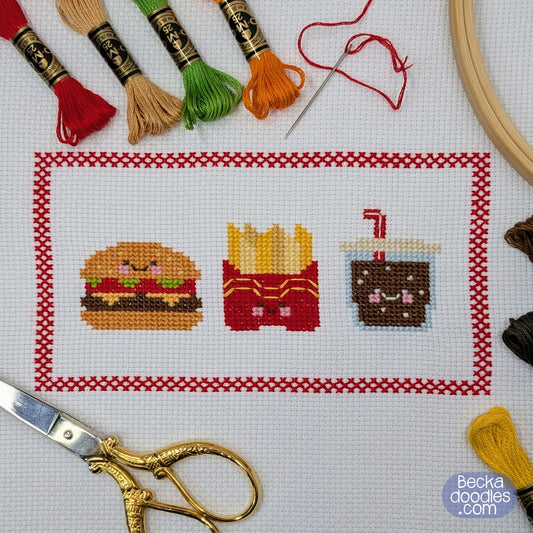 Cross-stitch design of a hamburger, french fries, and a drink, surrounded by embroidery tools including skeins of thread, a needle, scissors, and an embroidery hoop.