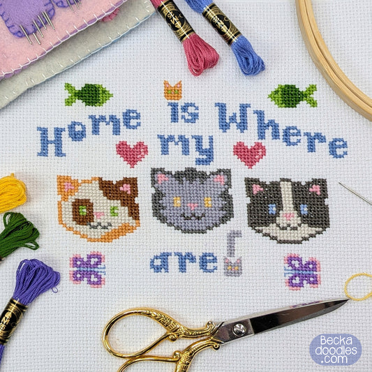 Cross stitch design with three cat faces and the phrase 'Home is Where My (cats) are!' stitched on white Aida with small fish, hearts, and butterflies added. Design is surrounded by embroidery tools.