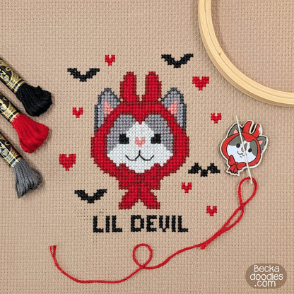 A cross-stitch design featuring a cute cartoon cat wearing a devil horns hat. Stitched text 'LIL DEVIL' below with small red hearts and black bats on light brown Aida fabric. A wooden hoop, needle and thread, and embroidery floss are arranged around the finished piece.