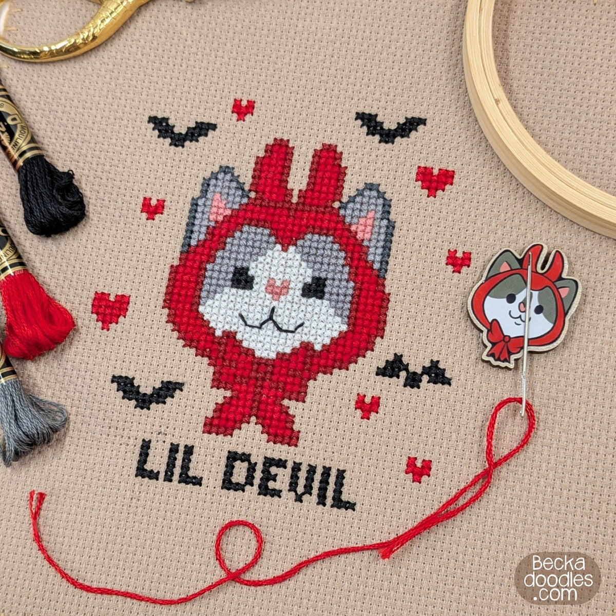 A close-up of a finished cross-stitch design featuring a cartoon cat wearing a red devil hat with the text 'LIL DEVIL' and small black bats and red hearts on light brown fabric. Surrounding props include embroidery floss, a wooden hoop, and a needle minder featuring the same artwork.
