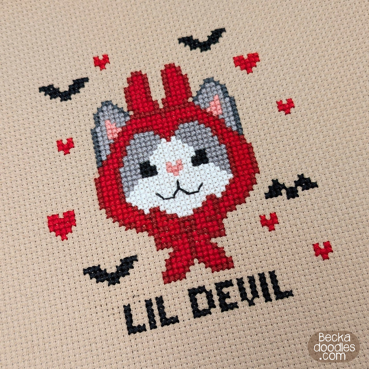 A close-up of a finished cross-stitch design featuring a cartoon cat in a red devil costume with black bats, red hearts, and the text 'LIL DEVIL' on a light brown fabric background. 