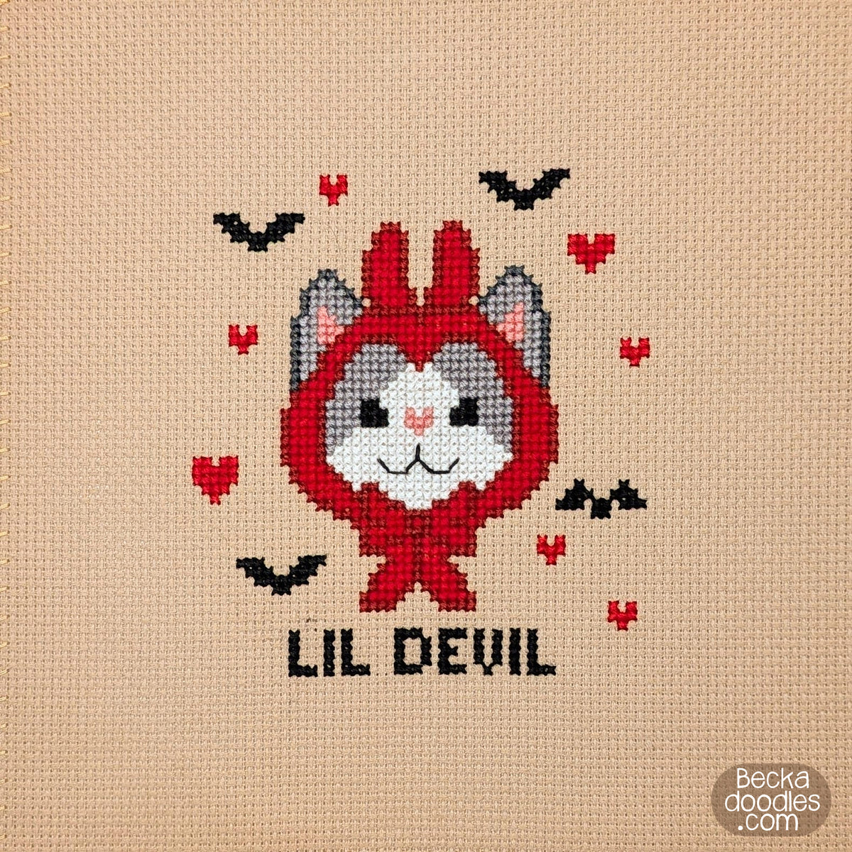 A cross-stitch design on beige fabric featuring a gray and white cat in a red devil costume with black bats and red hearts, with the text 'LIL DEVIL' stitched below.