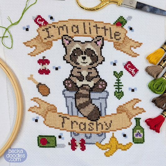 Cross-stitch design featuring a raccoon sitting on a trash can with banners reading "I'm a little Trashy" on a white fabric background. Surrounded by embroidery supplies.