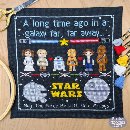 A square black cross-stitch sampler featuring a Star Wars themed pixel-art design with characters, lightsabers, the Death Star, and the quote 'May The Force Be With You, Always,' shown on a wooden surface with embroidery tools around it.