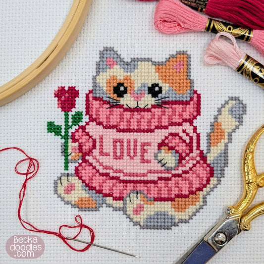 A completed cross-stitch design of a pretty calico cat wearing an pink "Love" sweater, surrounded by scissors, embroidery floss, a needle, a hoop on a white Adia fabric. 