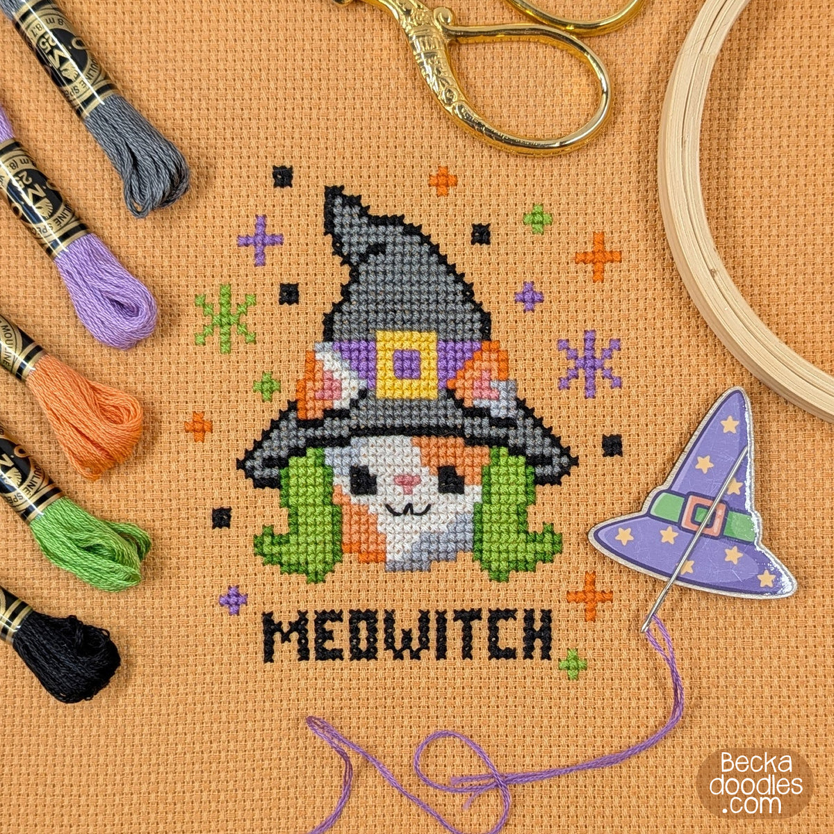 A cross-stitch design of a smiling calico cat wearing a witch hat and green hair with the word 'MEOWITCH' stitched below. Stitched on orange Aida cloth with embroidery floss, scissors, a wooden hoop, and a witch hat needle minder surrounding the design.
