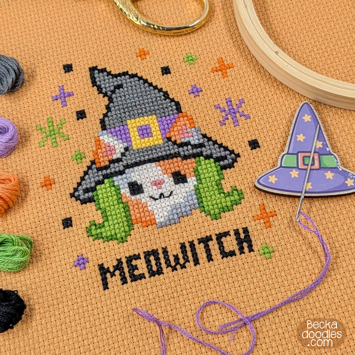 A close-up of a completed cross-stitch design featuring a cat dressed as a witch with the word “MEOWITCH” stitched below on orange Aida cloth. Embroidery threads, a wooden hoop, a purple witch hat needle minder, and a needle and thread surround the design.