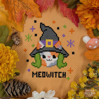 A cross-stitch design featuring a calico cat in a witch hat with green hair and  the word 'MEDWITCH' stitched below on orange Aida cloth. Design is surrounded by autumn leaves, flowers, pinecones, and a mushroom. 