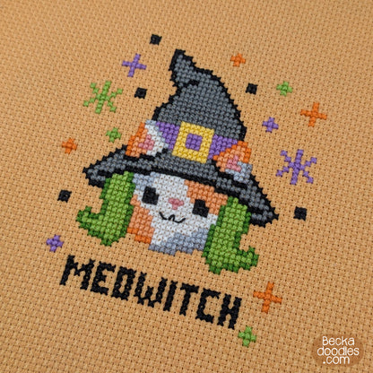 A close-up of a cross-stitch design featuring a cute calico cat dressed as a witch with a black hat, green hair, and the word 'MEDWITCH' stitched below on orange Aida cloth.