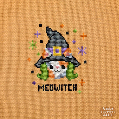 A cross-stitch design on orange fabric featuring a cute calico cat with a black witch hat and green hair, surrounded by small colorful stars, and the word "MEDWITCH" stitched below.