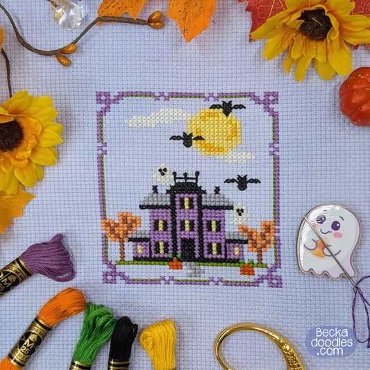 A finished cross-stitch design of a haunted purple mansion with a yellow moon, tiny bats and ghosts, autumn trees and a decorative purple border. Project is surrounded by embroidery supplies and fall foliage. 