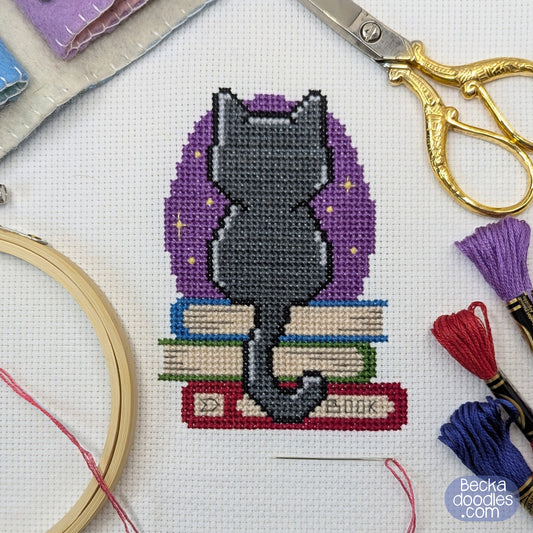 Cross-stitch pattern of a black cat on books with a purple star field surrounded by embroidery tools.