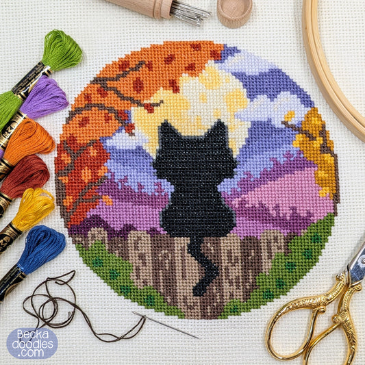 Cross-stitched design with a black cat silhouette in front of a full moon and a scenic landscape, surrounded by embroidery threads and tools.