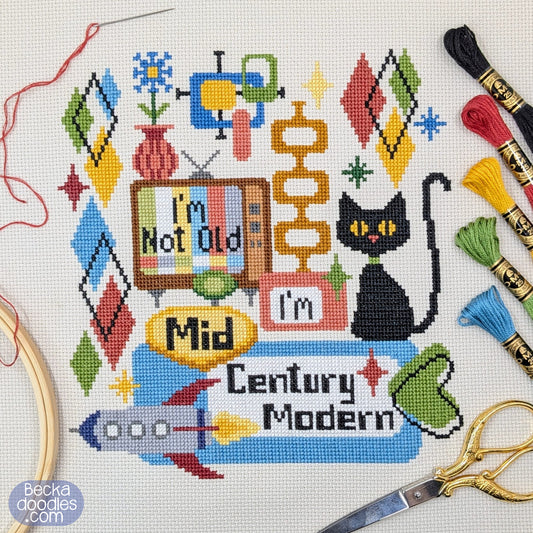 A colorful cross-stitch design on cream fabric featuring a black cat, retro TV, rocket, retro diamond shapes and starbursts, and the text 'I'm Not Old' and 'Mid Century Modern.' Scissors, thread skeins, and a wooden hoop are arranged around the stitched piece.