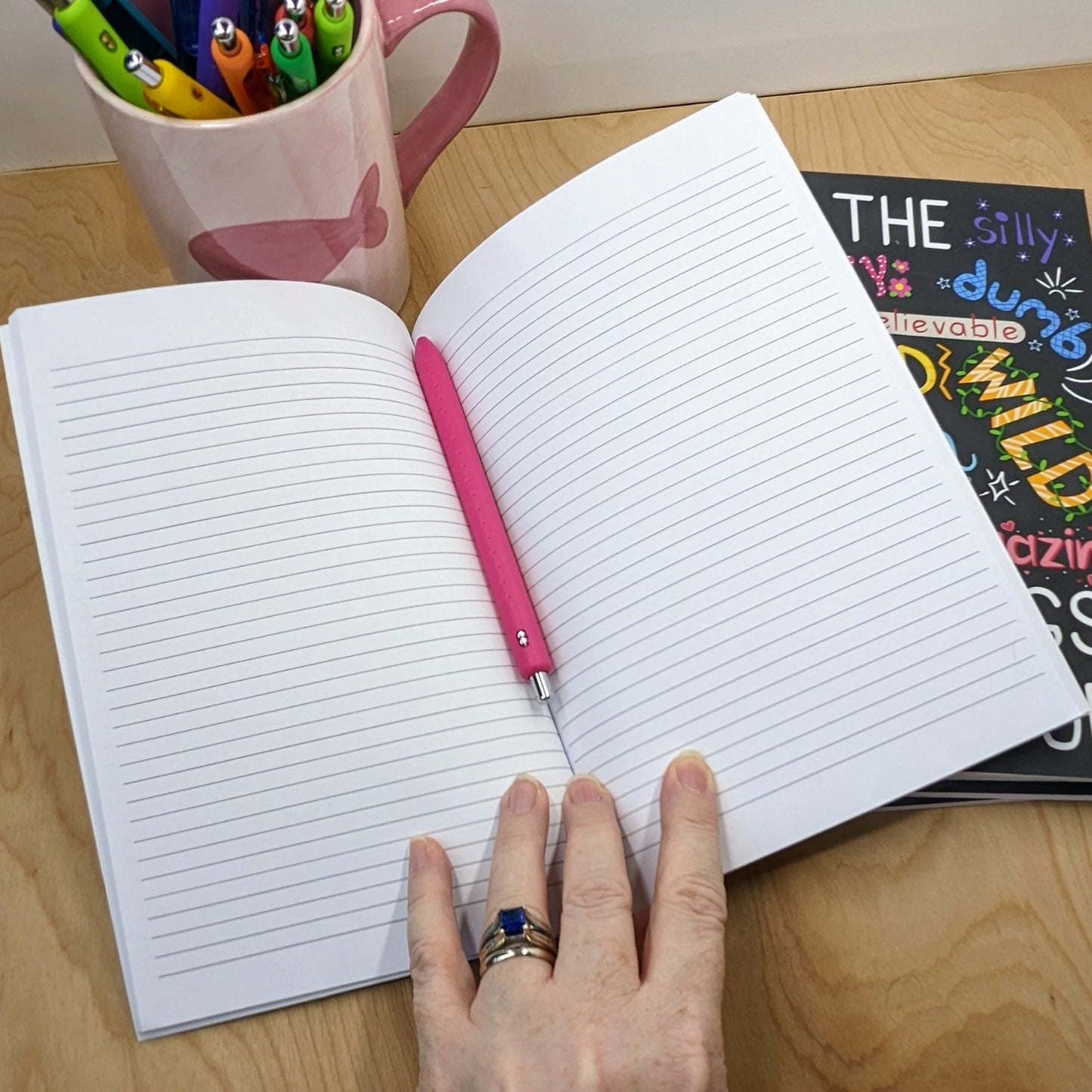 Open notebook with a pink pen on a wooden surface, next to a mug with pens and a book.