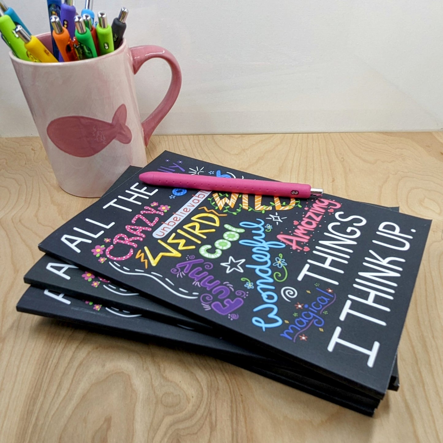Stack of notebooks with colorful text and a pink mug with pens on a wooden surface