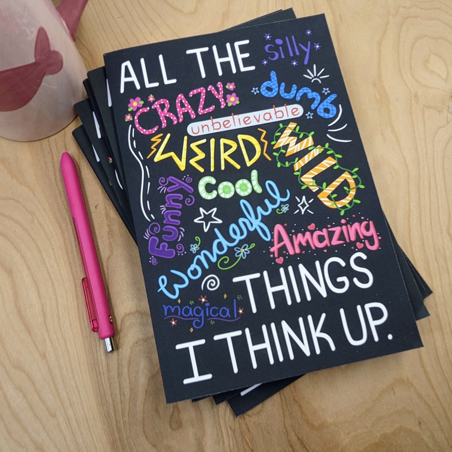 Black notebook with colorful text on a wooden surface with a pink pen.