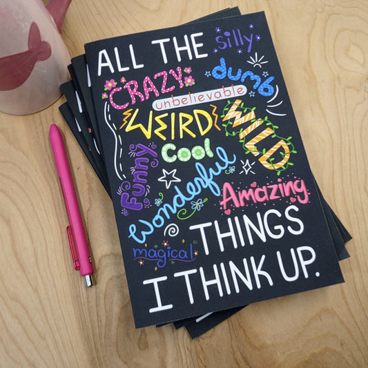 Black notebook with colorful text on a wooden surface with a pink pen.