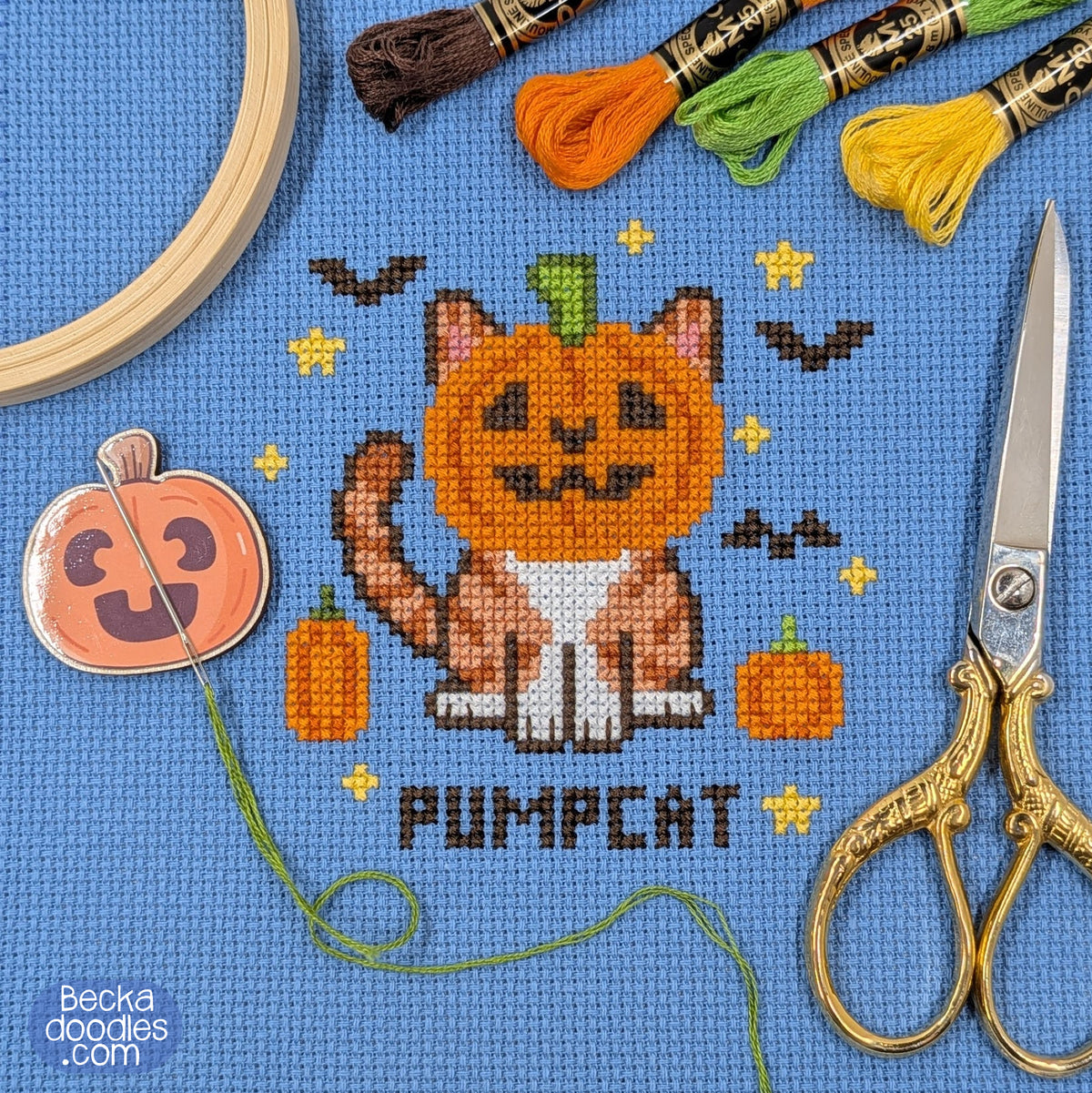 A cross-stitch design of a pumpkin-headed cat on blue Aida fabric with the word 'PUMPCAT' stitched below. Design is surrounded by a embroidery hoop, scissors, thread, and a pumpkin-shaped needle minder.