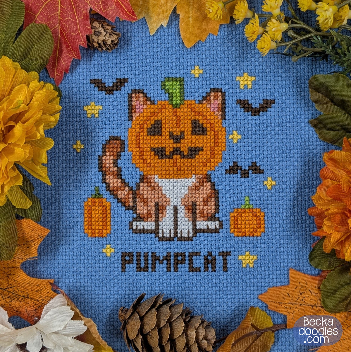 A cross-stitch design of a pumpkin-headed cat on a blue Aida cloth surrounded by autumn leaves, flowers, and pinecones. The text 'PUMPCAT' is stitched at the bottom of the design.
