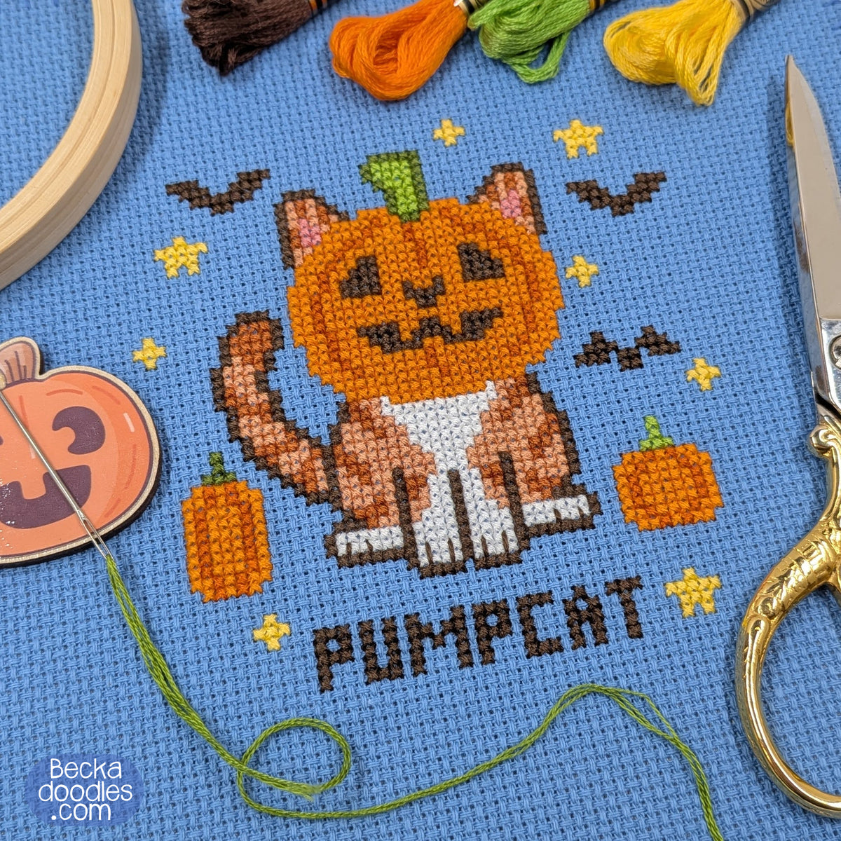A close-up of a cross-stitch design featuring a cat with a pumpkin head, surrounded by small pumpkins, bats, stars, and the word 'PUMPCAT' on a blue Aida cloth. Scissors, thread, a pumpkin needle minder, and a wooden hoop surround the design.