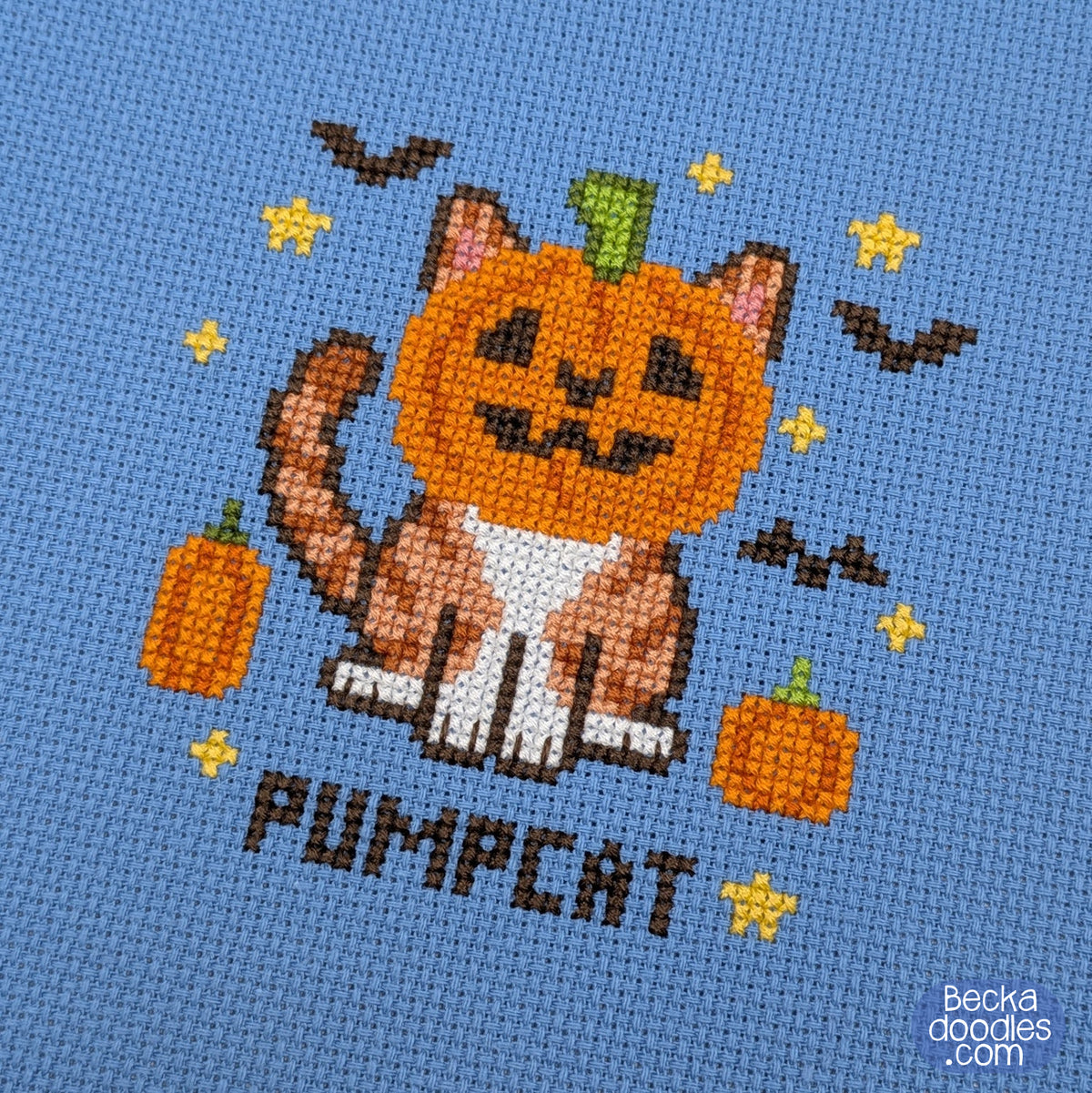 A close-up of a cross-stitch design featuring a cat with a pumpkin head, small orange pumpkins, yellow stars, bats, and the word "PUMPCAT" stitched on blue Aida cloth.