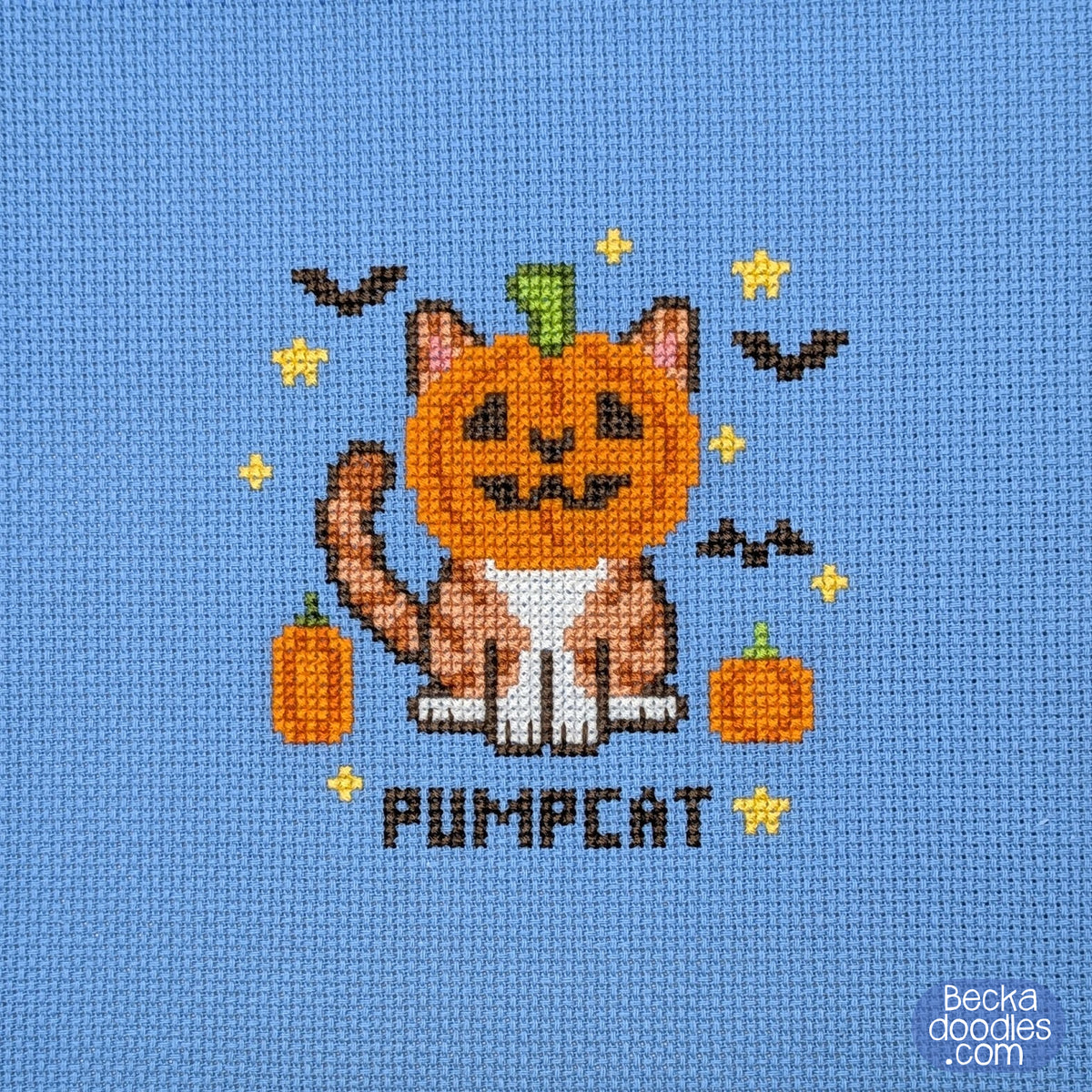 A cross-stitch design featuring a cat with a pumpkin head, small orange pumpkins, yellow stars, bats, and the word "PUMPCAT" stitched on blue Aida cloth.