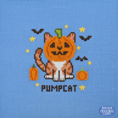 A cross-stitch design featuring a cat with a pumpkin head, small orange pumpkins, yellow stars, bats, and the word "PUMPCAT" stitched on blue Aida cloth.