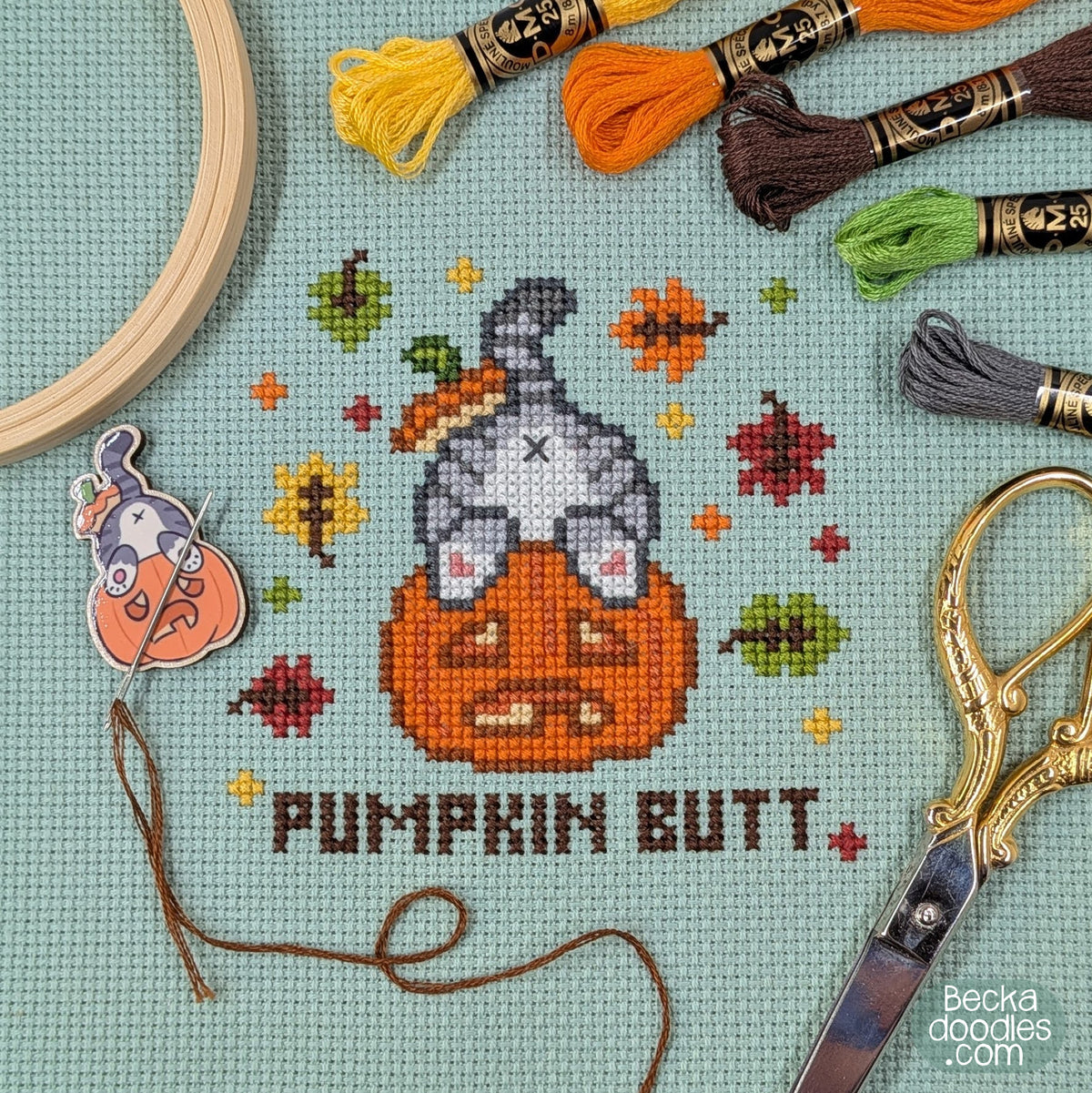 A cross-stitch design of a cat's butt sticking out of a pumpkin with the words 'PUMPKIN BUTT' below. Stitched on moss green Aida cloth surrounded by embroidery floss skeins, a wooden embroidery hoop, scissors, and a needle minder featuring the same design.