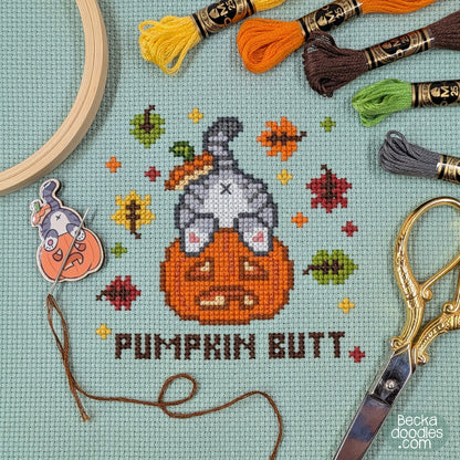 A cross-stitch design of a cat's butt sticking out of a pumpkin with the words 'PUMPKIN BUTT' below. Stitched on moss green Aida cloth surrounded by embroidery floss skeins, a wooden embroidery hoop, scissors, and a needle minder featuring the same design.