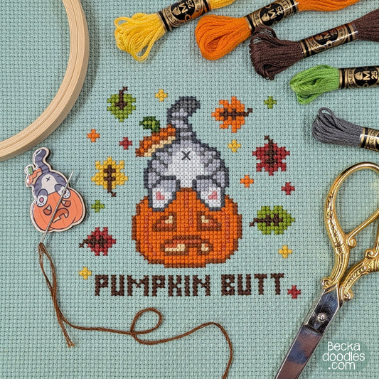 A cross-stitch design of a cat's butt sticking out of a pumpkin with the words 'PUMPKIN BUTT' below. Stitched on moss green Aida cloth surrounded by embroidery floss skeins, a wooden embroidery hoop, scissors, and a needle minder featuring the same design.