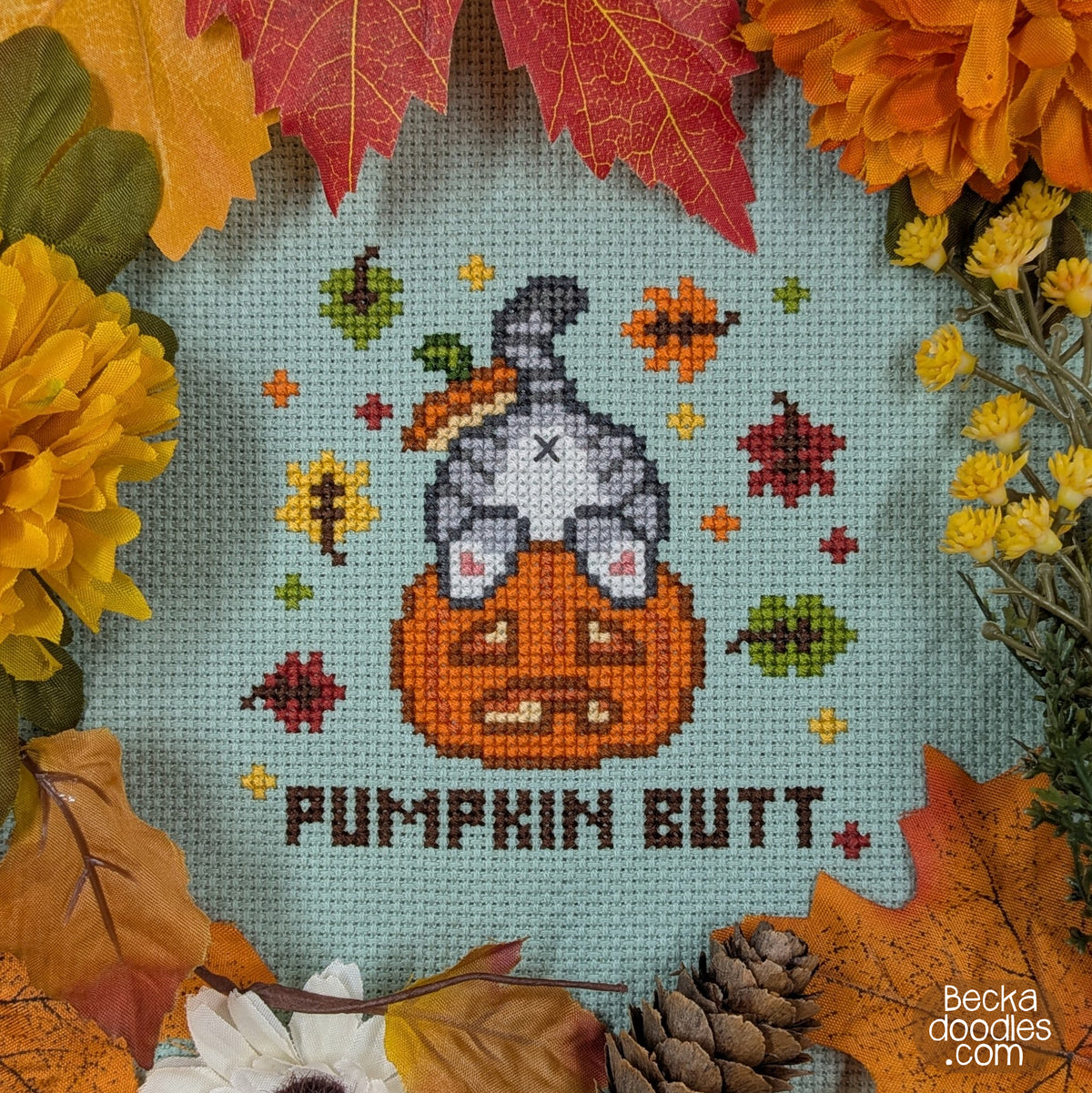 A cross-stitch design on moss green Aida cloth, featuring a gray cat's butt sticking out of a pumpkin surrounded by stitched fall leaves, and the text 'PUMPKIN BUTT.' The design is surrounded by autumn leaves, flowers, and a pinecone.