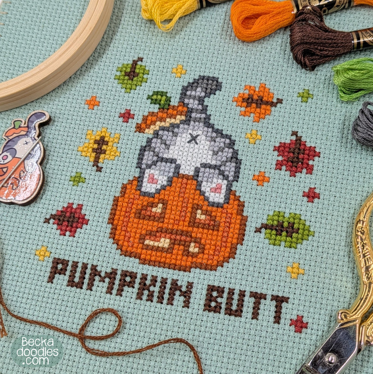 A close-up of a cross-stitch design featuring a gray cat's butt sticking out of a pumpkin with the text 'PUMPKIN BUTT' below. The stitching is on moss green Aida cloth, surrounded by embroidery tools and thread skeins, including a wooden hoop, yellow and orange thread, and a needle minder.