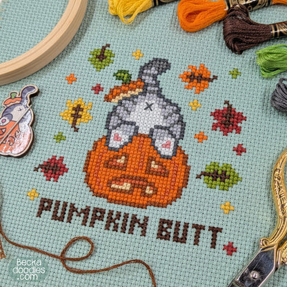 A close-up of a cross-stitch design featuring a gray cat's butt sticking out of a pumpkin with the text 'PUMPKIN BUTT' below. The stitching is on moss green Aida cloth, surrounded by embroidery tools and thread skeins, including a wooden hoop, yellow and orange thread, and a needle minder.