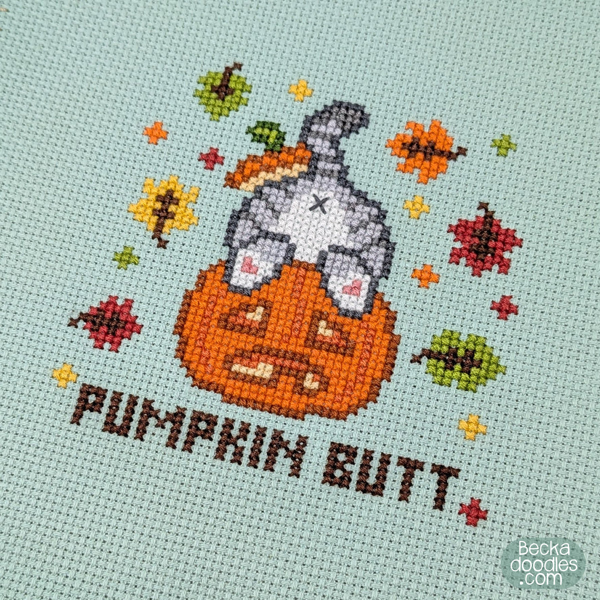 A close-up of a cross-stitch design featuring a gray cat's butt sticking out of a pumpkin with the text 'PUMPKIN BUTT' below and autumn leaves surrounding on moss green Aida cloth.