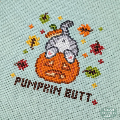 A close-up of a cross-stitch design featuring a gray cat's butt sticking out of a pumpkin with the text 'PUMPKIN BUTT' below and autumn leaves surrounding on moss green Aida cloth.