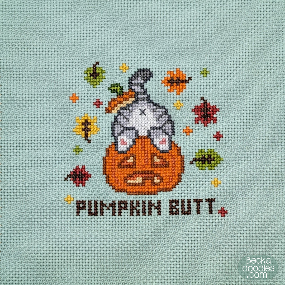 A close-up of a cross-stitch design featuring a gray cat's butt sticking out of a pumpkin with the text 'PUMPKIN BUTT' below and autumn leaves surrounding on moss green Aida cloth.