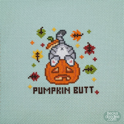 A close-up of a cross-stitch design featuring a gray cat's butt sticking out of a pumpkin with the text 'PUMPKIN BUTT' below and autumn leaves surrounding on moss green Aida cloth.