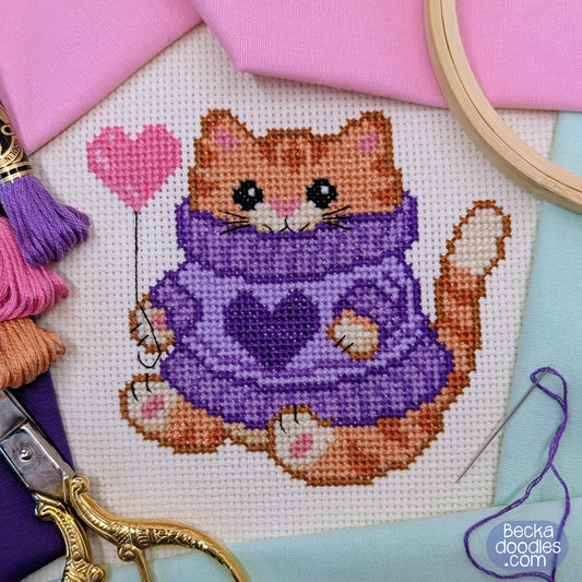 Cross-stitched cat in a purple sweater with a pink heart balloon, surrounded by embroidery tools and pastel fabrics.