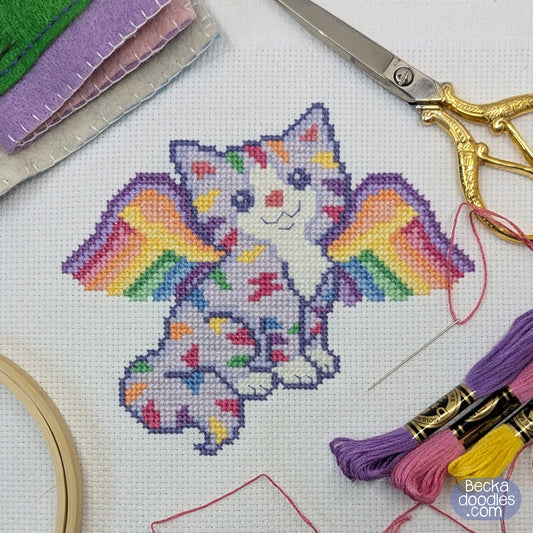 A cross-stitch design of a white cat with colorful rainbow wings, shown on white fabric with embroidery hoops, thread, scissors, and needles around it. BeckaDoodles branding is visible in the bottom right corner.