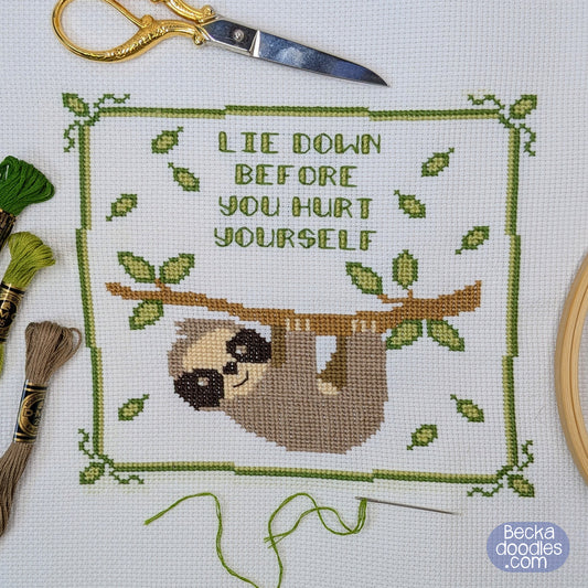Cross-stitch design of a brown sloth hanging from a branch with the text 'LIE DOWN BEFORE YOU HURT YOURSELF' inside a green leafy border displayed on white fabric surrounded by sewing supplies.